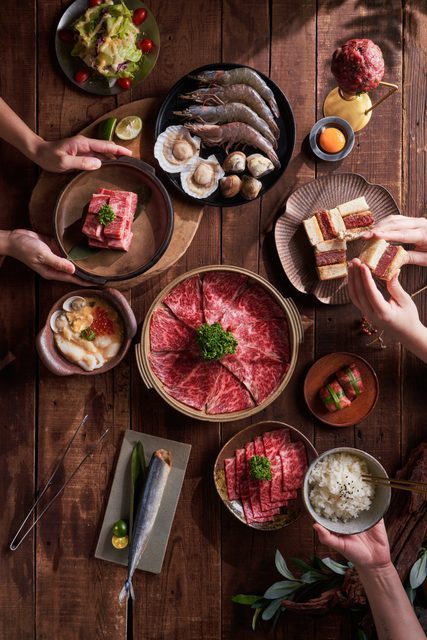 俯拍木桌上的肉品、配菜與餐具，多隻手從桌邊伸入