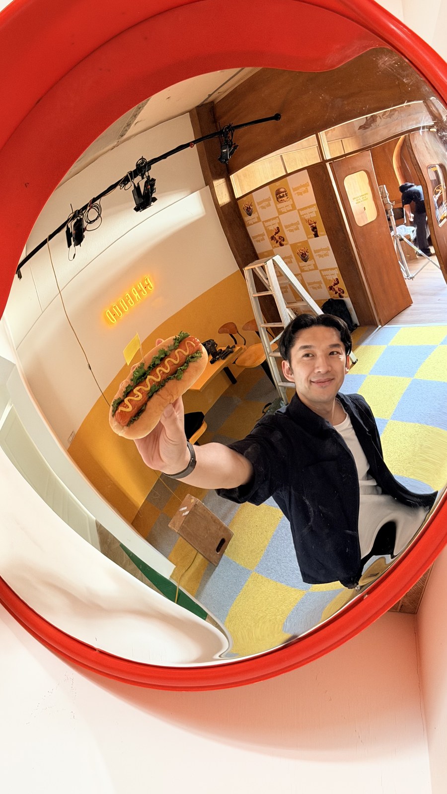 Nick 在紅色圓鏡前拿著熱狗堡,身後是色彩鮮明的店內空間