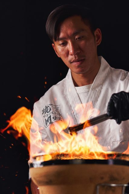 廚師在暗背景中操作炭爐，火焰照亮肉品與烤網